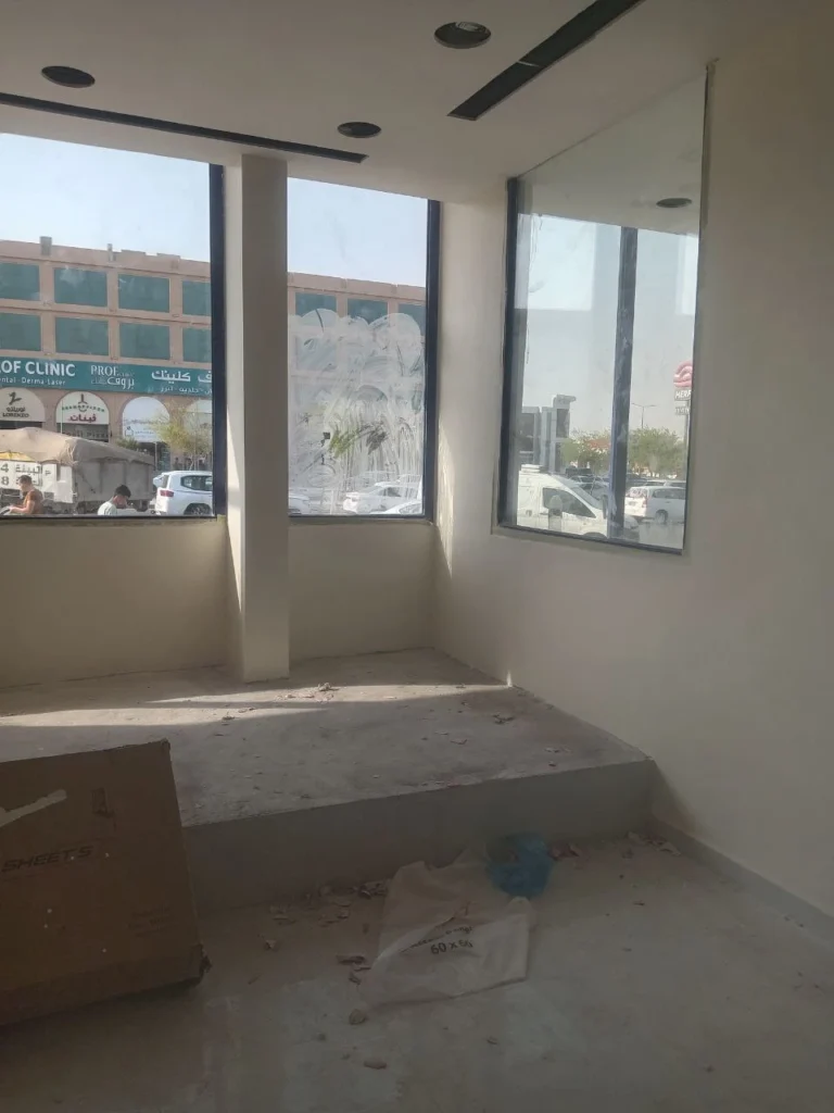 غرفة داخلية قيد التشطيب بجدران بيضاء ونوافذ كبيرة تطل على الشارع مع أرضية غير مكتملة وبعض مخلفات العمل